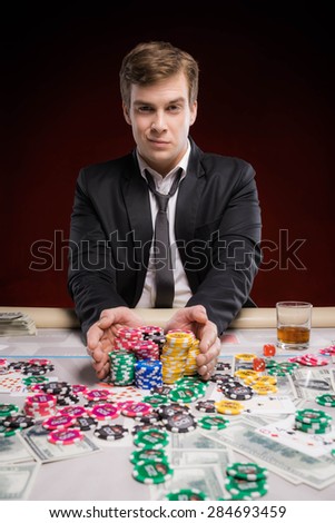 Young man with satisfied grin sitting at poker table, making bets and looking at camera. The chips, cards and money are on table