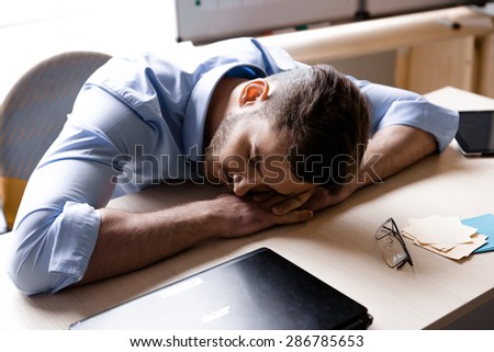 Young tired businessman sleeping near laptop in office full of folders for documents and office supplies