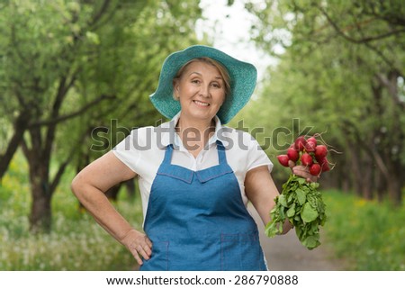 Good-looking smiling middle-aged woman-farmer holding freshly picked bunch of radishes and standing in green garden