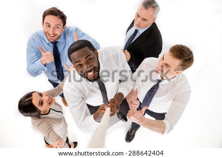 Top view of young smiling African-american businessman reaching up along rope. Other members of business team cheering him up. Isolated on white