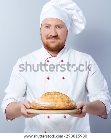 Portrait of positive young male chef in white uniform. Head-cook looking at camera and proposing freshly baked bread. Standing against grey background