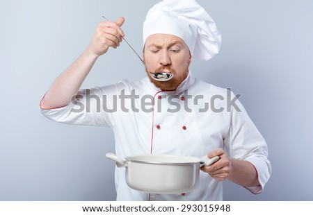 Portrait of young male chef in white uniform. Head-cook tasting food from saucepan and standing against grey background