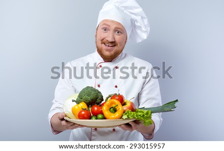 Portrait of positive young male chef in white uniform. Head-cook cheerfully smiling, looking at camera and proposing big plate of fresh vegetables. Standing against grey background