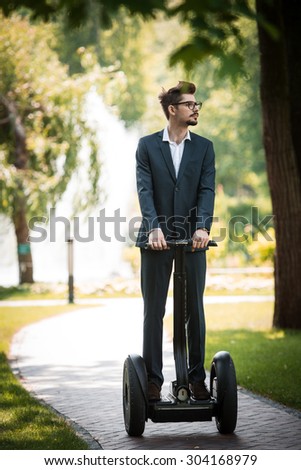 Portrait of handsome young businessman wearing suit. Man using segway and looking aside. Green alley as background