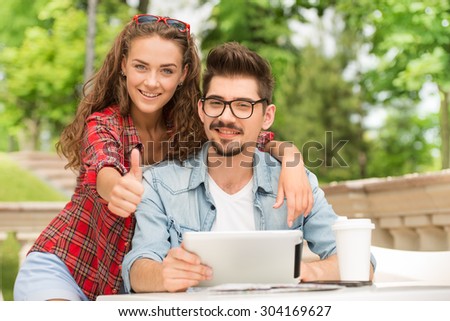 Portrait of beautiful young woman and handsome man on vacation. Girl and boy using tablet computer, hugging and smiling. Girl showing thumb up