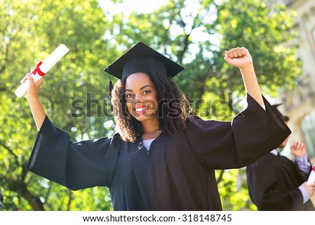 Young Afro American female student dressed in black graduation gown. Campus as a background. Girl cheerfully smiling with arms up, holding diploma and looking at camera