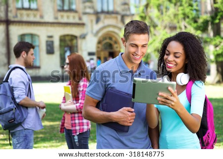 Photo of young group of students with backpacks and books. Campus as a background. Girl and boy using tablet computer. Students are on background