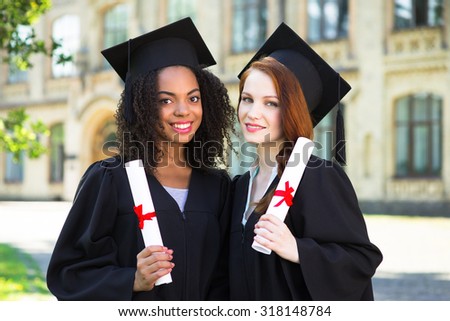 Young female students dressed in black graduation gown. Campus as a background. Girls smiling, holding diplomas and looking at camera