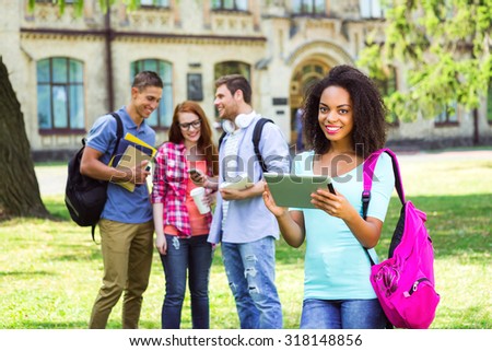 Photo of young group of students with backpacks and books. Campus as a background. Afro American girl smiling, using tablet computer and looking at camera. Students are on background