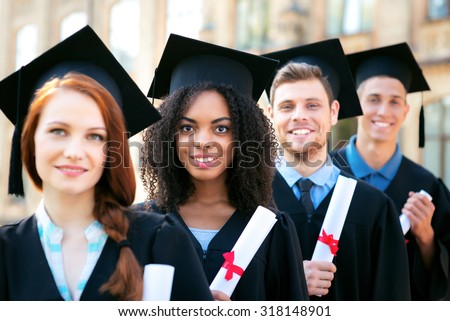Young students dressed in black graduation gown. Campus as a background. Students standing in row, smiling, holding diplomas and looking at camera. Focus on Afro American girl