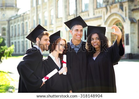 Young students dressed in black graduation gown. Campus as a background. Boys and girls smiling, hugging, holding diplomas and making photo on mobile phone