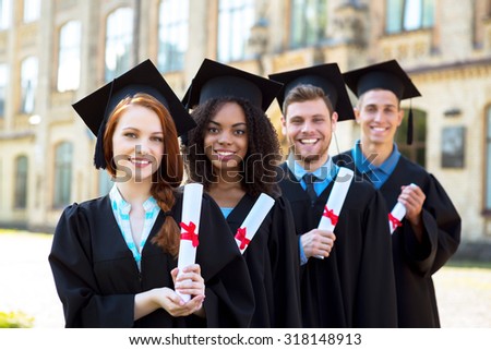 Young students dressed in black graduation gown. Campus as a background. Students standing in row, smiling, holding diplomas and looking at camera
