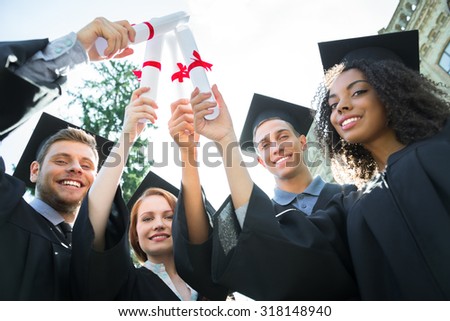 Young students dressed in black graduation gown. Campus as a background. Boys and girls cheerfully smiling, holding diplomas as swords and looking at camera. Bottom view photo