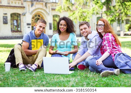 Young group of students with backpacks and books smiling, sitting on grass and using laptop. Campus as a background