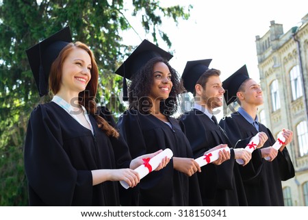 Young students dressed in black graduation gown. Campus as a background. Students standing in row, cheerfully smiling, holding diplomas and looking straight ahead