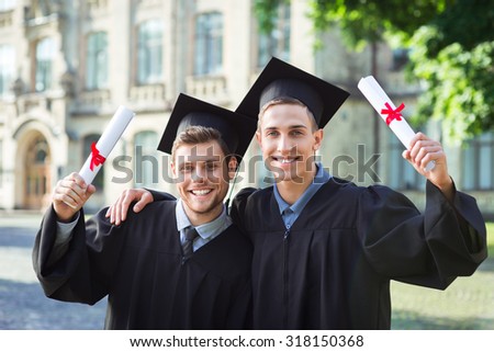 Young male students dressed in black graduation gown. Campus as a background. Boys cheerfully smiling, holding diplomas, embracing one another and looking at camera