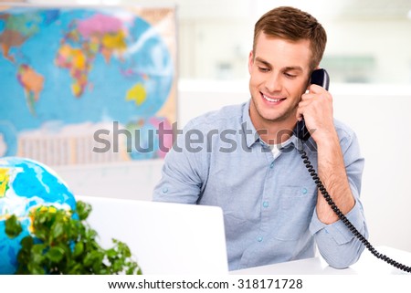 Photo of male travel agent. Young man talking on phone and smiling. Travel agency office interior with big world map
