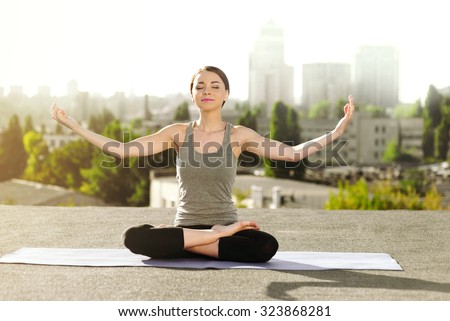 Young woman doing yoga on the mat on the roof, sitting in lotus posture and meditating