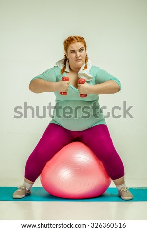 Funny picture of amusing, red haired, chubby woman on white background. Woman sitting on the ball and keep the dumbbells
