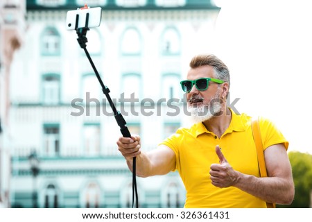 Portrait of stylish handsome adult man with beard standing outdoors. Man wearing glasses and yellow T-shirt, showing thumb up and making photo with selfie stick