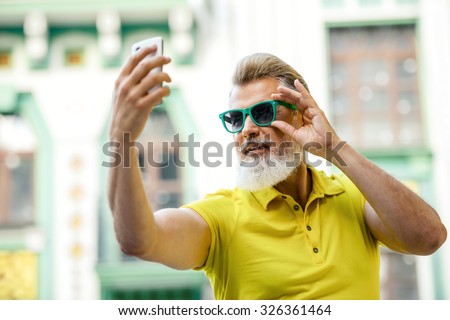 Portrait of stylish handsome adult man with beard standing outdoors. Man wearing glasses and yellow T-shirt, and making selfie photo with mobile phone
