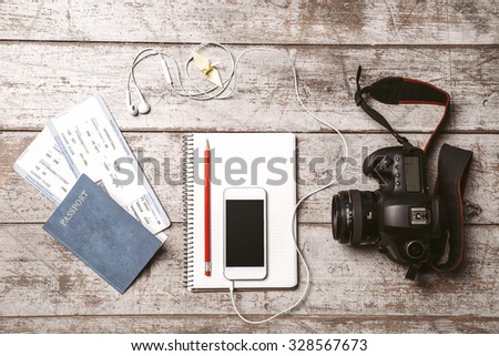 Top view photo of white mobile phone, professional camera, notebook, passport, tickets, pencil, origami bird and headphones. Objects are on light colored wooden floor