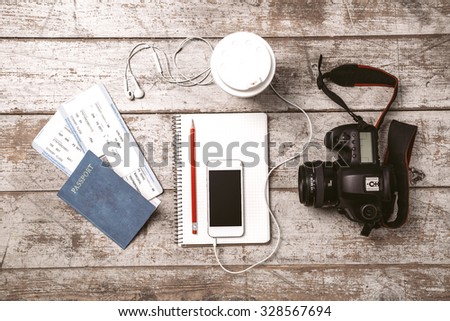 Top view photo of white mobile phone, professional camera, notebook, passport, tickets, pencil, cup and headphones. Objects are on light colored wooden floor