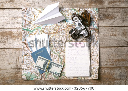 Top view photo of world map, white paper plane, passport, tickets, money, notebook and vintage camera. Objects are on light colored wooden floor