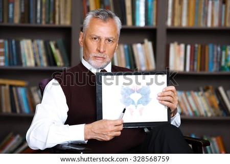 Portrait of male psychologist. Psychologist standing near big cupboard with books in psychologist office and holding Rorschach paintings