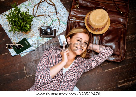 Top view photo of beautiful blonde girl lying on wooden floor. Young woman smiling, holding credit card and looking at camera. Passport, tickets, vintage camera, hat and map are on floor