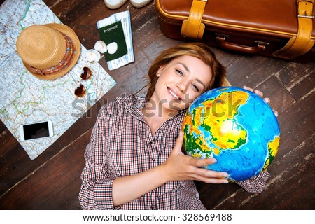 Top view photo of beautiful blonde girl lying on wooden floor. Young woman smiling, holding globe and looking at camera. Passport, tickets, mobile phone, hat, suitcase and map are on floor