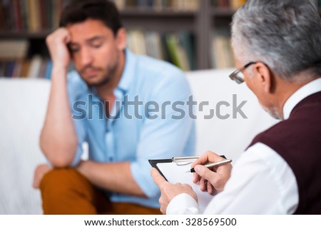 Sad young man talking with psychologist. Psychologist taking notes. There are many books in psychologist office