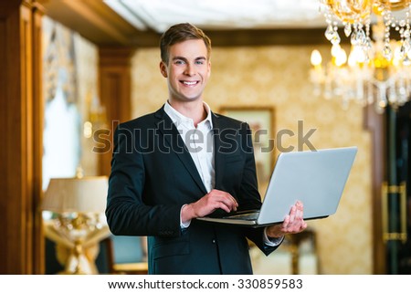 Photo of businessman in expensive hotel. Young smiling businessman wearing suit, standing in nice hotel room, using laptop and looking at camera