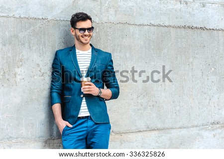 Portrait of stylish handsome young man with bristle standing outdoors. Man wearing jacket and shirt. Smiling man with sunglasses holding cup of coffee and leaning against wall
