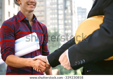 Portrait of engineer and builder. Men with helmet and blueprints standing near new unfinished construction. Men shaking hands after deal