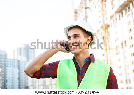 Portrait of builder in uniform. Young man with helmet standing near new unfinished construction and talking on mobile phone