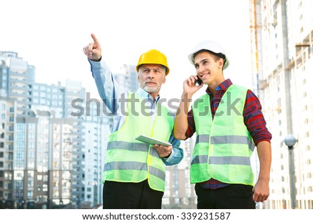 Portrait of contractor and builder in uniforms. Men with helmets standing near new unfinished construction and using tablet computer with mobile phone
