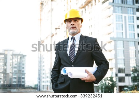Business portrait of contractor and developer. Aged businessman with helmet standing near his new construction and holding blueprints