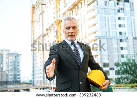 Business portrait of contractor and developer. Aged businessman with helmet standing near his new construction. Man proposing hand to shake and looking at camera
