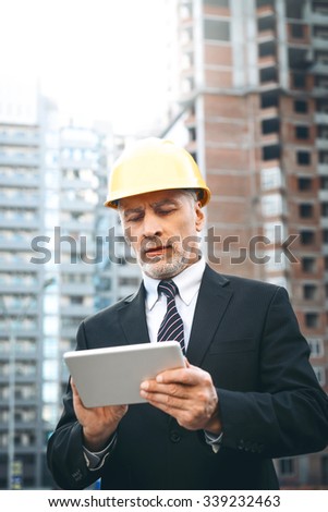 Business portrait of contractor and developer. Aged businessman with helmet standing near his new construction and using tablet computer