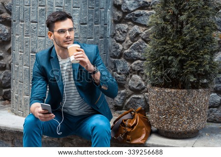 Portrait of stylish handsome young man with bristle standing outdoors. Man wearing jacket and watch. Man with glasses holding cup of coffee and using mobile phone