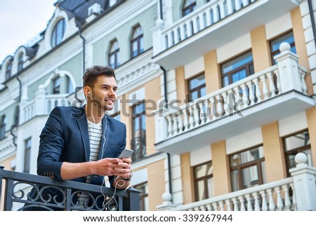 Portrait of stylish handsome young man with bristle standing outdoors. Man wearing jacket and shirt. Smiling man listening to music with mobile phone and leaning on parapet