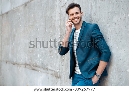 Portrait of stylish handsome young man with bristle standing outdoors and leaning on wall. Man wearing jacket and shirt. Smiling man using mobile phone