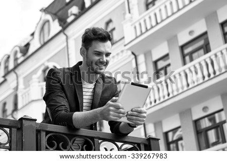Portrait of stylish handsome young man with bristle standing outdoors. Man wearing jacket and shirt. Smiling man using tablet computer. Black and white photo