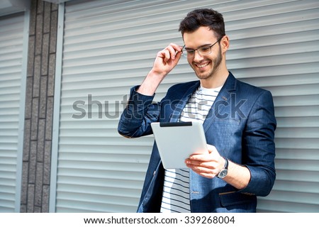 Portrait of stylish handsome young man with bristle standing outdoors. Man wearing jacket, glasses and shirt. Smiling man using tablet computer