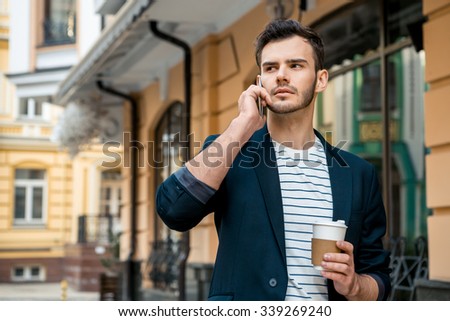 Portrait of stylish handsome young man with bristle standing outdoors. Man wearing jacket and shirt. Young man talking on mobile phone and holding cup of coffee