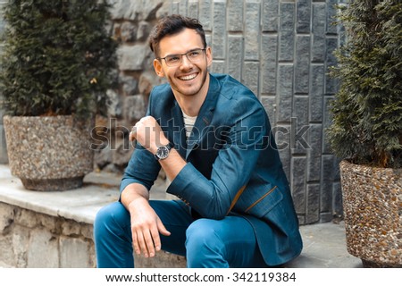 Portrait of stylish handsome young man with bristle standing outdoors. Man wearing jacket and watch. Man with glasses cheerfully smiling