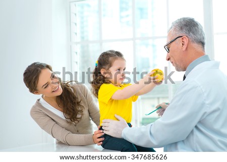 Portrait of aged doctor, little patient and her mother. They are in hospital office with big window. Medico diagnosing little girl