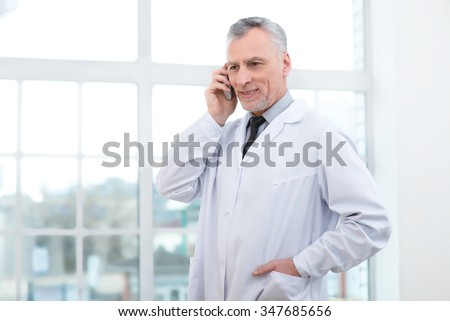 Portrait of aged doctor wearing lab coat. Doctor in years standing in hospital office with big window. Medico smiling and using mobile phone