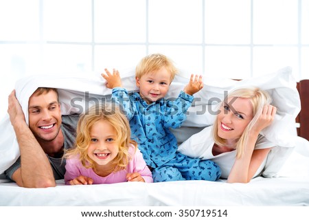 Photo of loving family of four lying on white bed in morning. Family hiding under blanket
